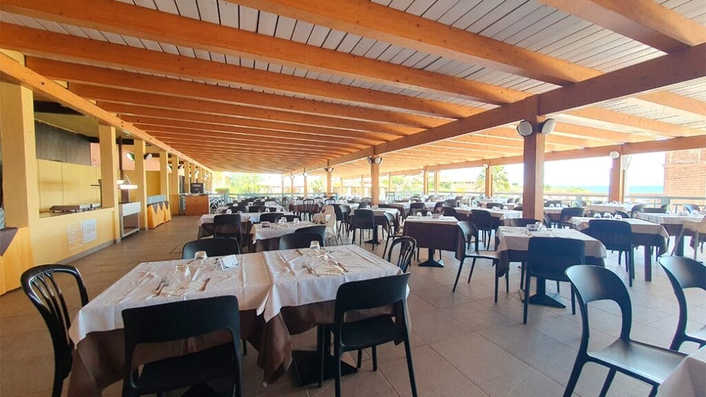 Ampio ristorante con travi in legno, tavoli apparecchiati, sedie nere e grandi finestre che lasciano entrare la luce del sole: il luogo perfetto per gustare una Bozza automatica in un'atmosfera calda e invitante.