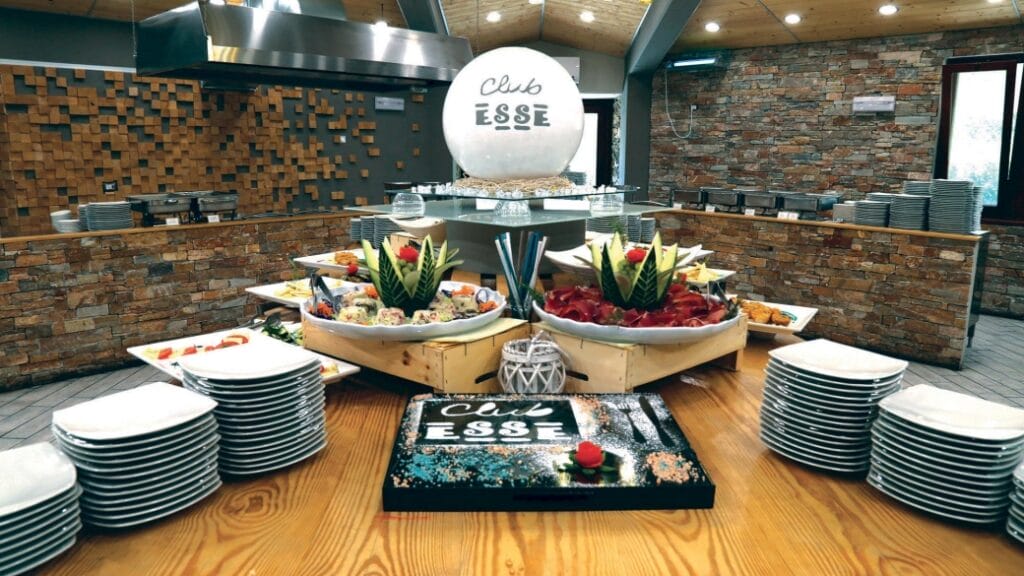 Tavolo da buffet con piatti, antipasti assortiti, intagli di frutta e un'insegna del Club Esse in una sala da pranzo rustica, il tutto allestito alla perfezione da Bozza automatica.