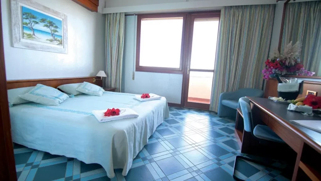 Camera d'albergo al Club Esse Roccaruja di Stintino con letto matrimoniale, decorazioni blu, scrivania, fiori e luce del sole che filtra dalle ampie finestre.