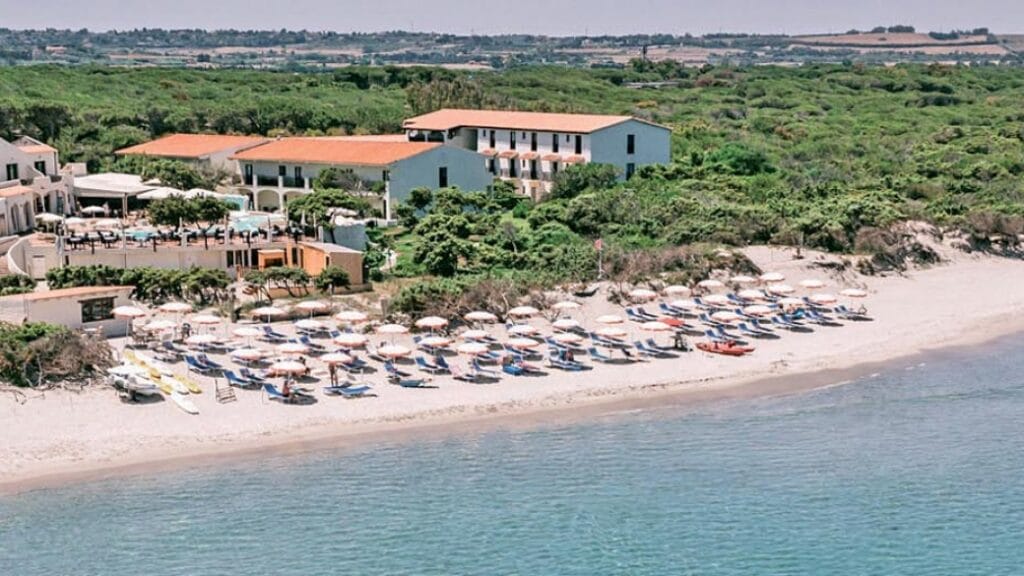 Vista aerea di un resort sulla spiaggia con ombrelloni e lettini in riva al mare, circondato dal verde: uno scenario invitante per una rilassante vacanza alla Bozza automatica.