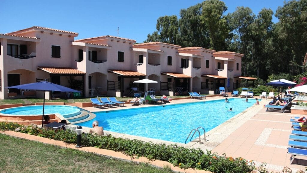 File di ville color beige si affacciano su una soleggiata piscina all'aperto, dotata di lettini e persone che si rilassano a bordo acqua, creando un'atmosfera tranquilla, perfetta per rilassarsi.