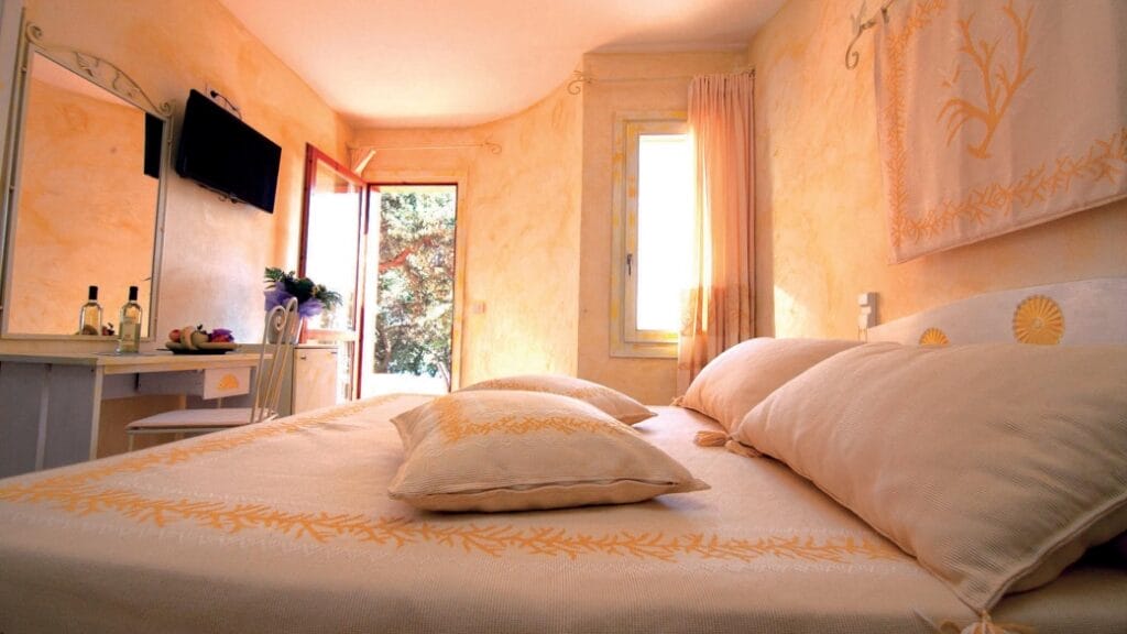 Accogliente camera da letto con letto matrimoniale, cuscini, mobiletto per toeletta, TV, porta aperta e luce naturale che entra a fiotti: perfetta per rilassarsi o per guardare i propri programmi preferiti con la comodità della Bozza automatica.