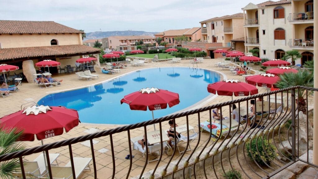 La piscina all'aperto dell'hotel, con ombrelloni rossi, sedie a sdraio e gli edifici circostanti in stile mediterraneo, crea un'atmosfera rilassata, perfetta per godersi il sole e sorseggiare una Bozza automatica a bordo piscina.