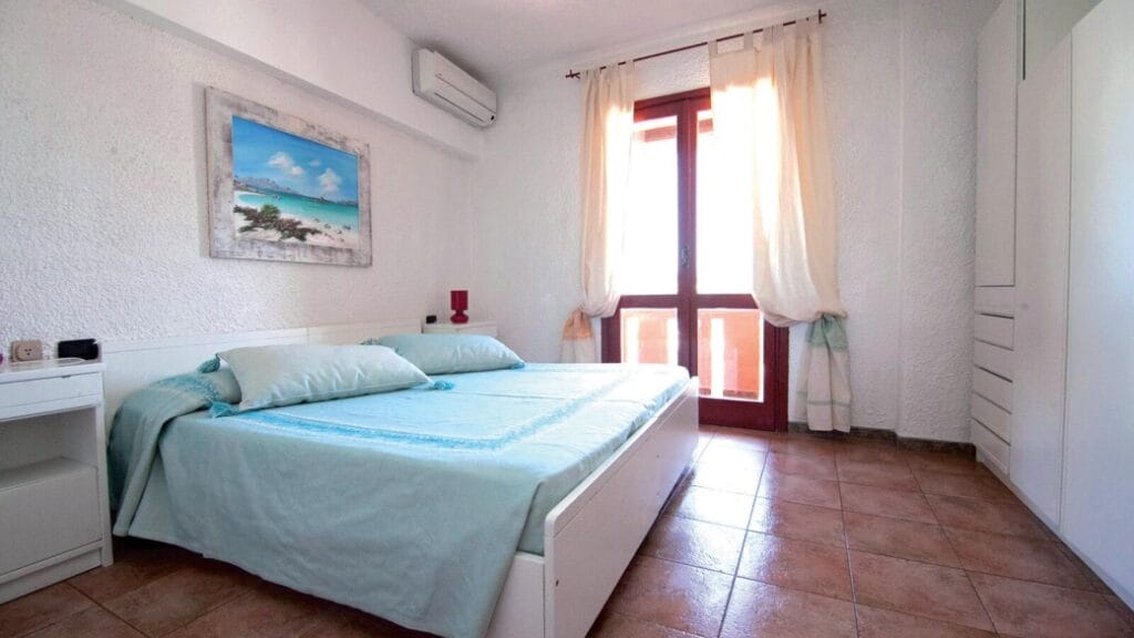 La luminosa camera da letto, dotata di letto matrimoniale, biancheria azzurra, quadri alle pareti e una grande finestra con tende trasparenti, crea uno spazio tranquillo e accogliente, perfetto per rilassarsi o lavorare sulla vostra ultima Bozza automatica.