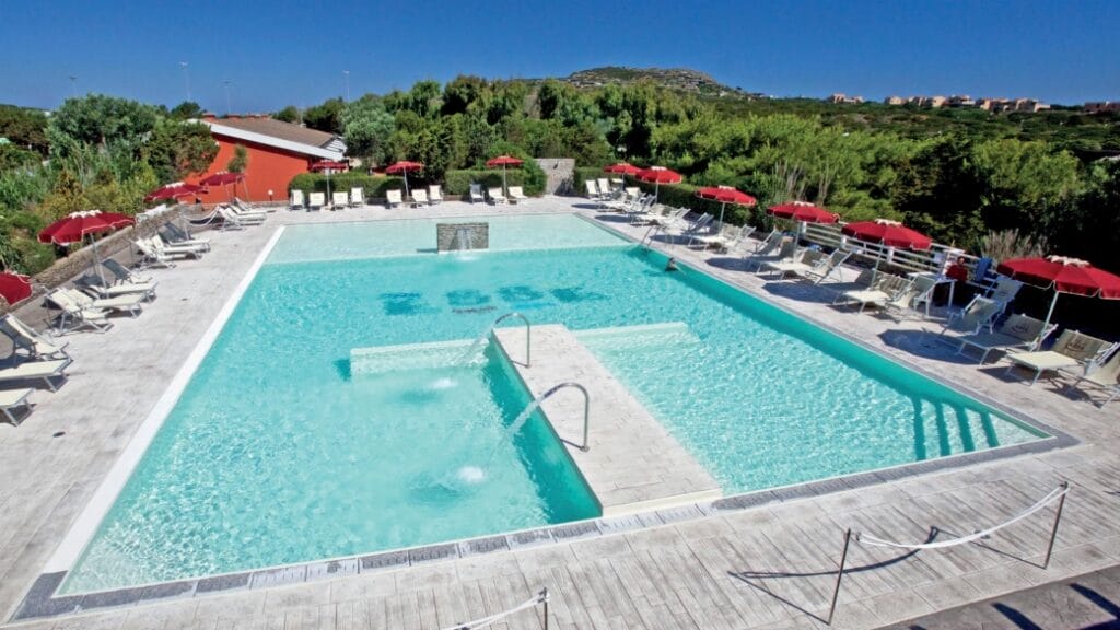 Ampia piscina esterna con lettini e ombrelloni rossi, circondata da alberi e cielo azzurro: perfetta per pomeriggi di relax. Godetevi la tranquillità con il nostro sistema automatico Bozza, che garantisce un ambiente sicuro e pulito.