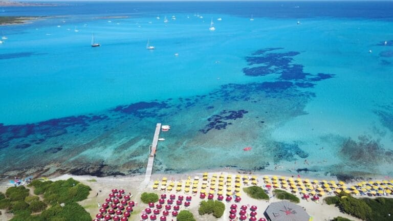 Vista aerea di una spiaggia con ombrelloni colorati, acqua azzurra e cristallina e diverse barche che galleggiano al largo, immortalata in una splendida bozza automatica.
