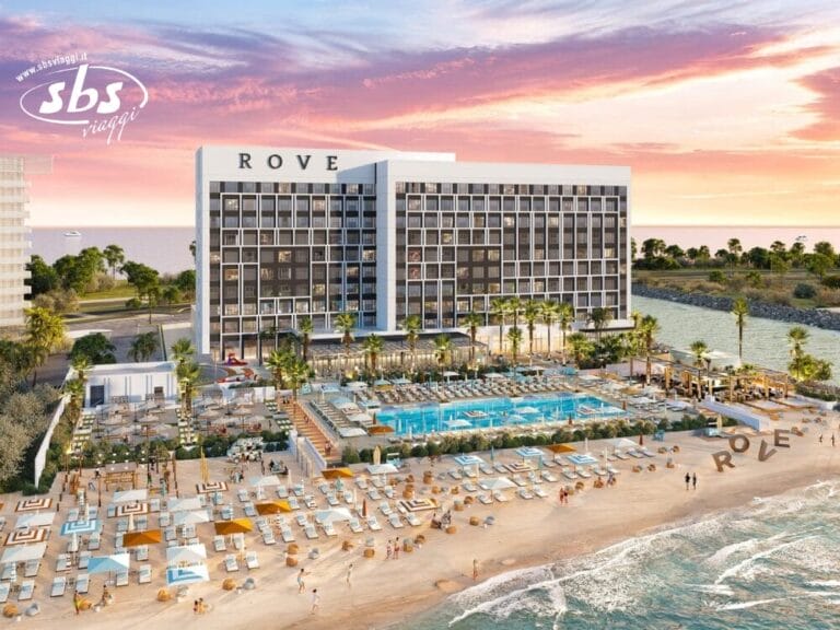 Un moderno hotel sulla spiaggia con una grande piscina, palme e lettini prendisole con vista sull'oceano al tramonto.