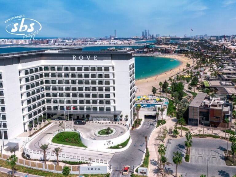 Vista aerea dell'hotel Rove vicino a una spiaggia sabbiosa con acqua azzurra e cristallina e lo skyline della città sullo sfondo, con una scena vivace che ricorda la Bozza automatica.