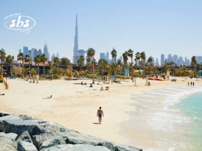 Una persona cammina su una spiaggia sabbiosa con palme e sullo sfondo lo skyline di Dubai, incluso il Burj Khalifa, immortalando il momento perfetto per una Bozza automatica.