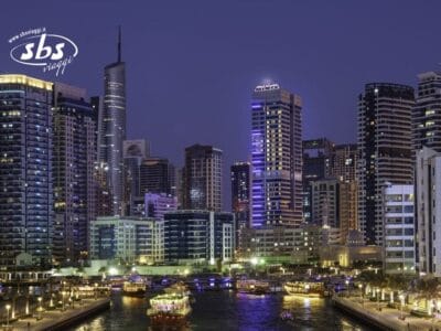 Lo skyline della città al tramonto, con i grattacieli illuminati e le barche che scivolano lungo il canale, evoca il fascino di Stella di Mare, con il logo "sbs viaggi" nell'angolo.