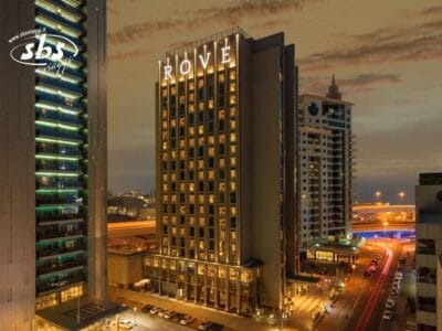 Vista notturna dell'edificio illuminato dell'hotel Rove, una presenza sorprendente tra le luci della città e gli alti edifici vicini, catturata nei minimi dettagli con Bozza automatica.