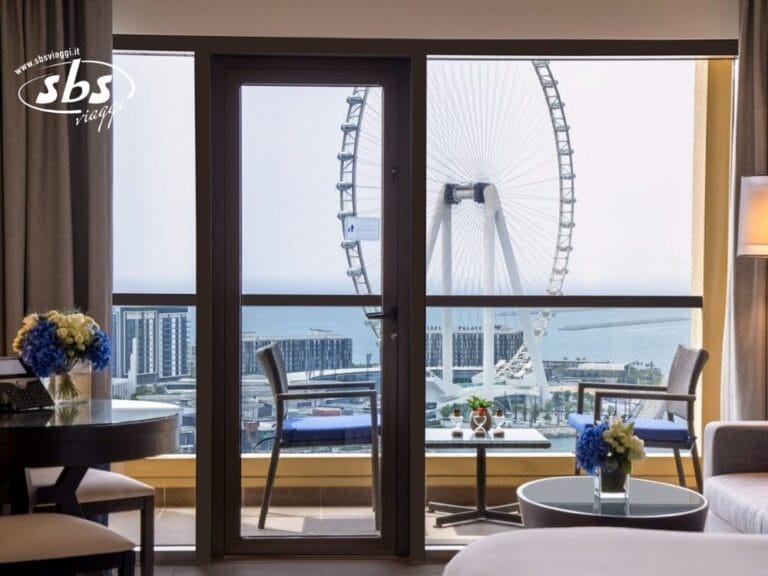 Camera d'albergo con balcone con vista sulla ruota panoramica gigante in riva al mare, fiori sul tavolo e servizi Bozza automatica per rendere il vostro soggiorno ancora più piacevole.