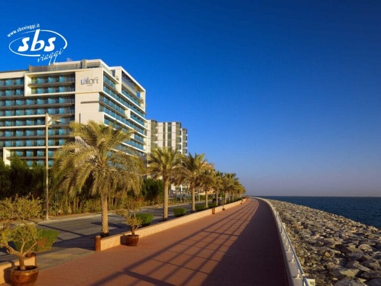 Un moderno hotel e palme costeggiano un soleggiato lungomare, caratterizzato da una costa rocciosa sotto un cielo azzurro e terso: un luogo invitante, perfetto per preparare la tua prossima Bozza automatica.