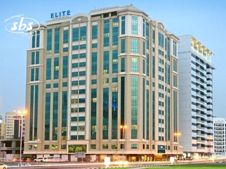 Un alto edificio alberghiero denominato "Elite", con finestre in vetro e finiture beige, sorge accanto a una strada cittadina al tramonto e il suo aspetto moderno è impreziosito da elementi di design Bozza Automatica.