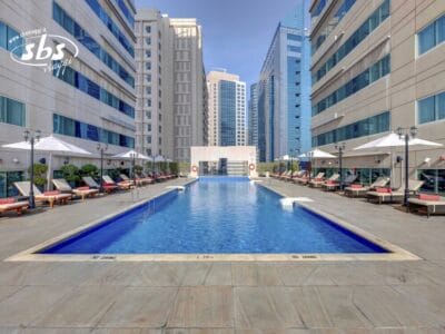 Piscina esterna rettangolare tra alti edifici moderni, con sedie a sdraio e ombrelloni su entrambi i lati: un'oasi invitante nel paesaggio urbano.