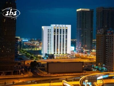 L'edificio dell'hotel Rove illuminato di notte, circondato dalle luci della città e dagli alti grattacieli, si erge come una vivace bozza automatica nello skyline urbano.
