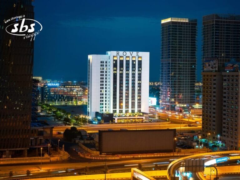 L'edificio dell'hotel Rove illuminato di notte, circondato dalle luci della città e dagli alti grattacieli, si erge come una vivace bozza automatica nello skyline urbano.
