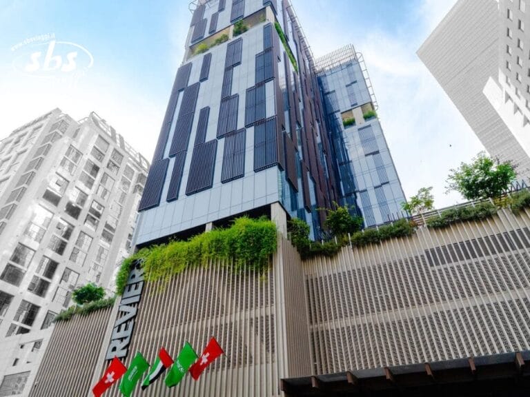 Moderno edificio alto con Bozza automatica, caratterizzato da vegetazione e bandiere sulla facciata, visto da un'angolazione bassa contro il cielo.