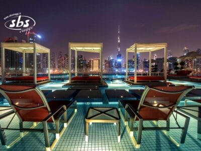 Le cabine a bordo piscina e le sedie a sdraio si affacciano sullo skyline della città di notte, con i grattacieli illuminati sullo sfondo, creando un'atmosfera Bozza automatica di eleganza urbana e relax.