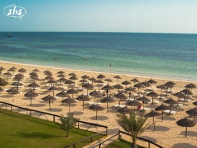 File di ombrelloni di paglia e sedie a sdraio costeggiano una spiaggia sabbiosa accanto al mare calmo e azzurro, sotto un cielo limpido; alcune persone nuotano e si rilassano vicino alla riva nella splendida Tunisia.