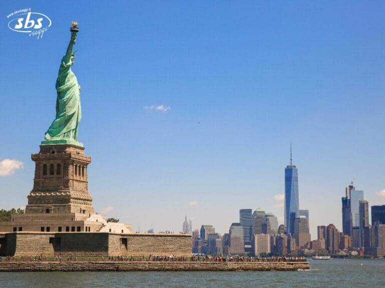 La Statua della Libertà si erge su Liberty Island con lo skyline di Manhattan, incluso il One World Trade Center, sullo sfondo: una tappa obbligata per ogni accompagnatore italiano che stia pianificando un'avventura di Ferragosto a New York sotto il limpido cielo azzurro.