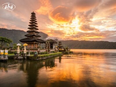 Un tempio tradizionale balinese sorge sulle rive di un lago calmo al tramonto, con nuvole spettacolari e la calda luce del sole che si riflette sull'acqua.