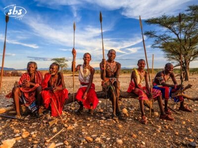 Sei persone vestite in abiti tradizionali siedono su un tronco all'aperto, impugnando delle lance sotto un cielo azzurro con nuvole sparse: una scena vibrante di scoperta culturale in Kenya.