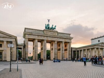 L'immagine mostra la Porta di Brandeburgo a Berlino al tramonto, con persone radunate attorno alla piazza e un cartello stradale con la scritta "Pariser Platz" in primo piano.