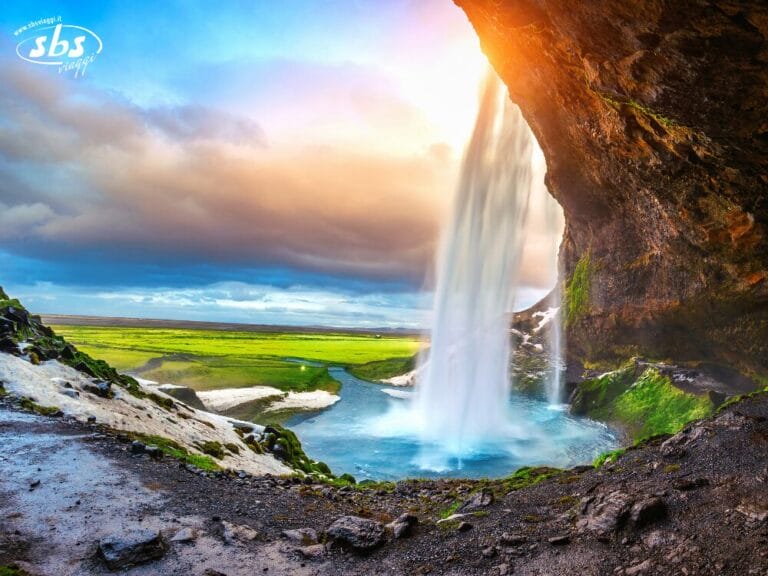 Una splendida cascata sgorga da una parete rocciosa e si riversa in un lussureggiante paesaggio verde sotto un cielo colorato al tramonto, catturando la bellezza selvaggia dell'Islanda che ricorda la terra nuova.