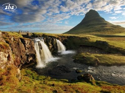 Una pittoresca cascata scorre in un paesaggio erboso con una ripida montagna conica sullo sfondo e un cielo azzurro, catturando la bellezza mozzafiato dei Meravigliosi Altopiani.