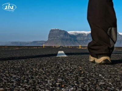 Una persona è in piedi su una strada deserta in Islanda, con una montagna sullo sfondo e un cielo azzurro e limpido: perfetto per un'avventura con il Classic Circle Tour.