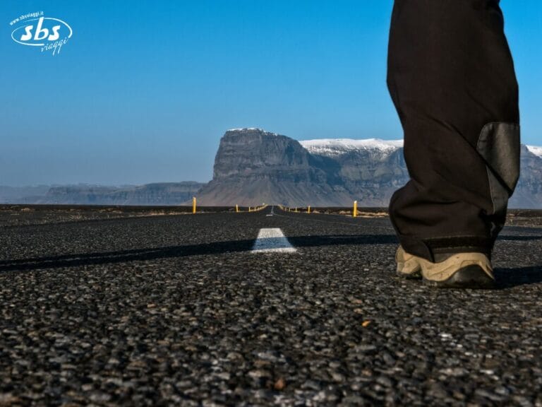 Una persona è in piedi su una strada deserta in Islanda, con una montagna sullo sfondo e un cielo azzurro e limpido: perfetto per un'avventura con il Classic Circle Tour.