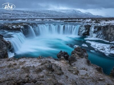 Ampia cascata che si riversa in una piscina turchese in mezzo a un terreno roccioso e innevato sotto un cielo nuvoloso; sullo sfondo colline innevate.