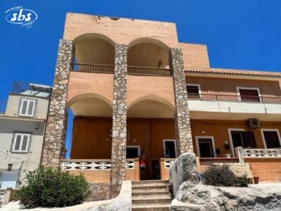 Case Vacanza Lampedusa, edificio a due piani con archi in pietra, pareti color cuoio e un balcone, con sfondo un cielo azzurro e terso.