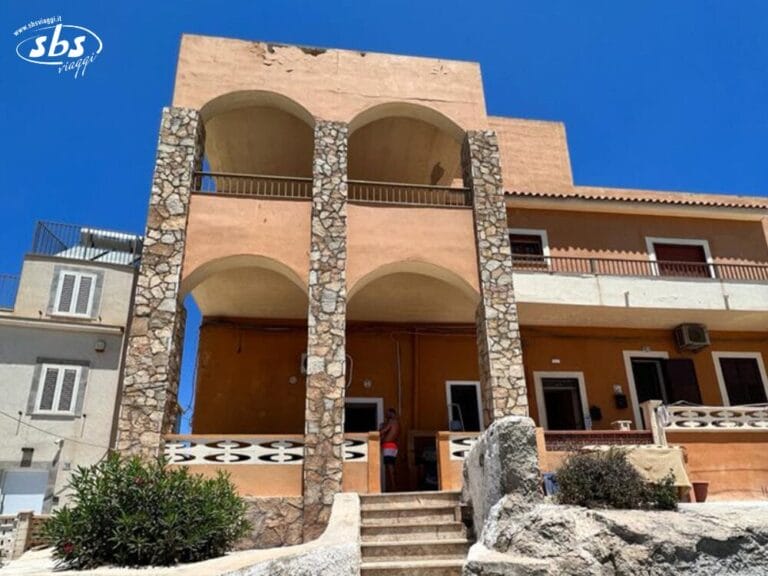 Case Vacanza Lampedusa, edificio a due piani con archi in pietra, pareti color cuoio e un balcone, con sfondo un cielo azzurro e terso.