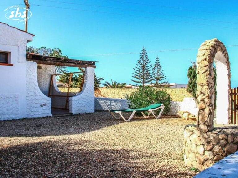 Affascinante casa in pietra bianca con ingresso ad arco, lettino prendisole e cortile con ghiaia sotto un cielo azzurro e limpido: perfetta per il tuo soggiorno al Residence Lampedusa, la scelta migliore per l'alloggio a Lampedusa.