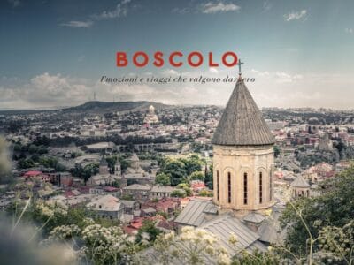 Un paesaggio urbano della Georgia con il campanile di una chiesa in primo piano e colline che ricordano l'Armenia sullo sfondo, sotto un cielo parzialmente nuvoloso.