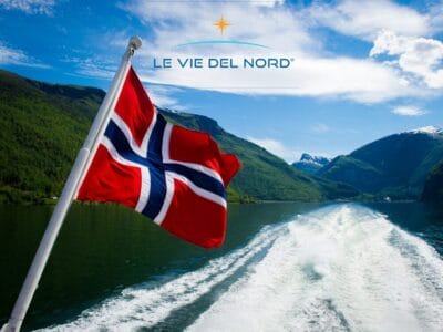 Bandiera norvegese su una barca con fiordi meravigliosi, montagne e cielo azzurro; sopra il testo "Le Vie Del Nord": un perfetto tour con accompagnatore dall'Italia.