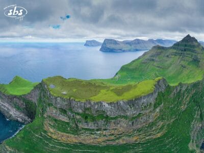 Vista aerea delle ripide scogliere verdeggianti delle Isole Faroe che si affacciano sull'oceano, con isole lontane sotto un cielo nuvoloso. Alcuni piccoli edifici sono visibili sull'altopiano erboso.