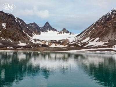 Le montagne innevate delle Isole Svalbard si riflettono in un lago calmo e turchese sotto un cielo nuvoloso, con chiazze di neve visibili sui pendii accidentati e rocciosi.
