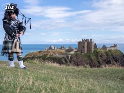 Un uomo in abiti tradizionali scozzesi suona la cornamusa su una scogliera erbosa, con le rovine di un castello e il mare sullo sfondo, sotto un cielo parzialmente nuvoloso.