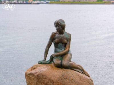 Una statua in bronzo di una sirena è seduta su una roccia vicino all'acqua, con la costa sfocata sullo sfondo.