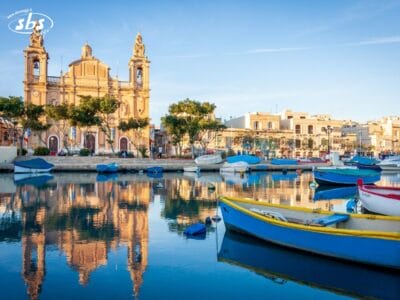 Barche colorate galleggiano sulle acque calme di un porto, con una chiesa storica e degli edifici che si riflettono nell'acqua sotto un cielo azzurro e limpido.