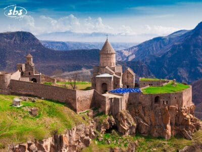 Un complesso monastico in pietra sorge su una scogliera rocciosa circondata da montagne, con una tenda blu e bianca vicino all'ingresso.