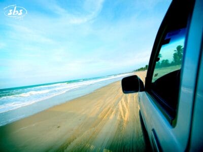 Un veicolo Fly & Drive percorre una spiaggia sabbiosa in riva all'oceano sotto un cielo azzurro, con le tracce degli pneumatici visibili sulla sabbia.