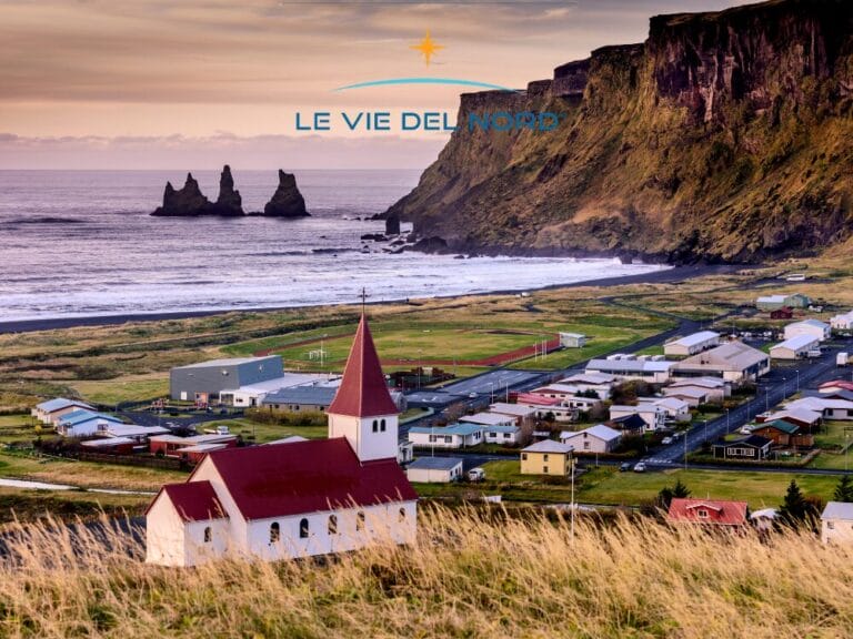 Villaggio costiero del Sud Islanda con chiesa dai tetti rossi, scogliere e faraglioni al tramonto sulle Isole Vestman; in alto il logo "Le Vie del Nord".