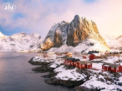 Lungo una costa rocciosa, con grandi montagne sullo sfondo, sotto un cielo parzialmente nuvoloso, si trovano delle cabine rosse coperte di neve.