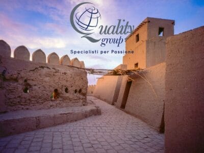 Antica fortezza in mattoni di fango in Uzbekistan al tramonto, con il logo "Quality Group, Specialisti per Passione" sovrapposto: un'esperienza Grantour indimenticabile.