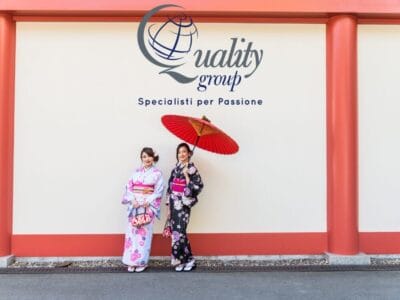 Due donne in kimono sono in piedi vicino a un muro, una delle quali regge un ombrello rosso, sotto il logo "Quality Group", che cattura perfettamente l'eleganza del Giappone e ispira i sogni del tuo prossimo Tour Giappone.