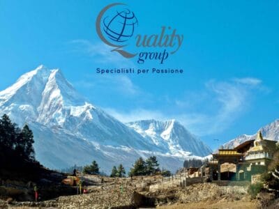 Montagna innevata e villaggio con il logo "Quality group"; il testo italiano nel cielo azzurro sopra evidenzia Valle degli Dèi e Tour Nepal.